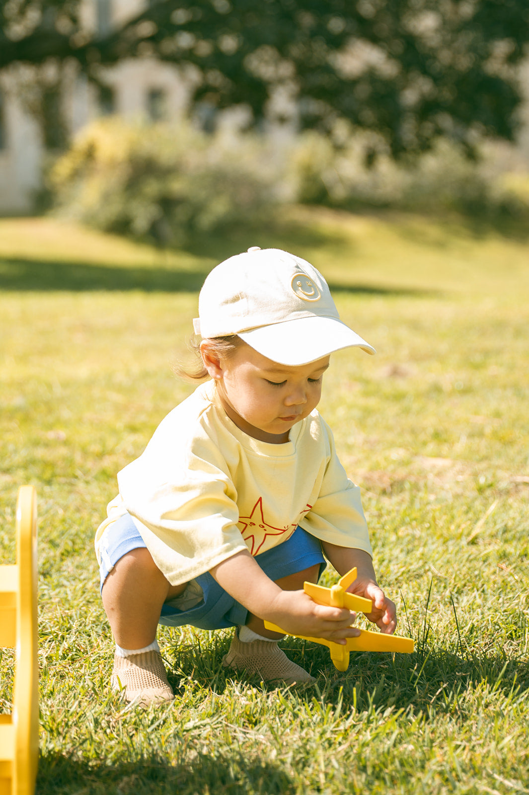 A toddler crouching on a grassy lawn, wearing a cream cap, a cream t-shirt with a red star graphic, and blue shorts, brown sohls while playing with a yellow toy airplane.