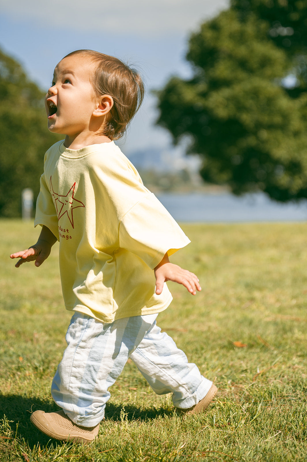 A toddler walking outdoors on a grassy field, wearing a pale yellow oversized T-shirt with a red star graphic and light blue pants with sohls toddler shoes