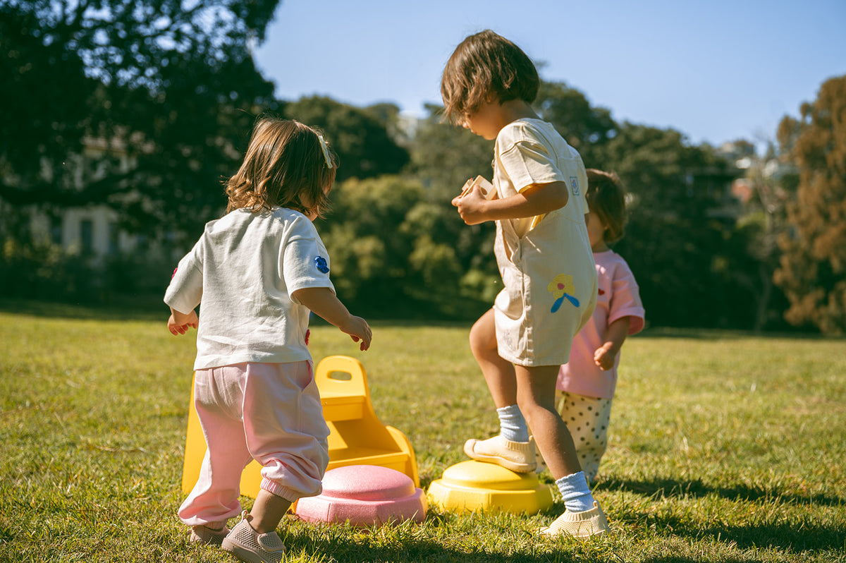 Three young children playing outdoors on a sunny grassy lawn with a yellow and pink plastic toy in the foreground and trees in the background.